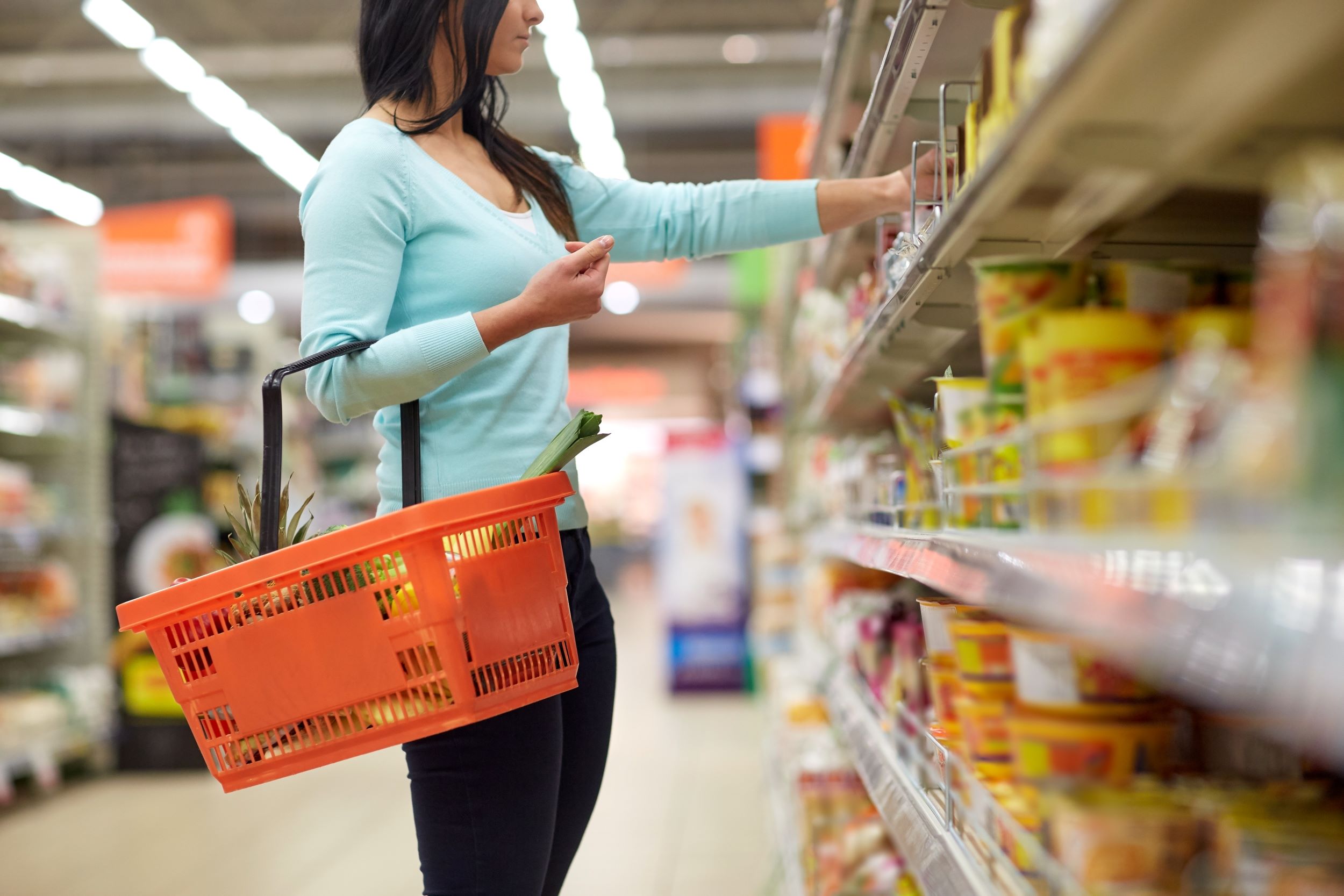 A woman with a basket in a supermarket picking up an item from a shelf